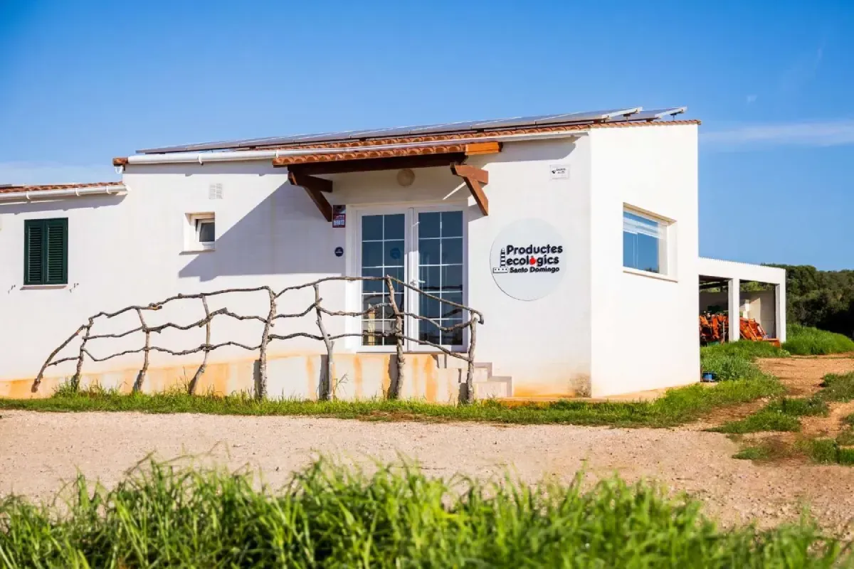 Pequeña casa rural blanca con porche y muro de piedra, situada junto a un campo de hierba bajo un cielo soleado.