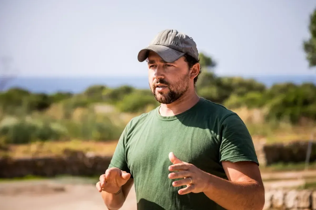 Hombre con camisa y gorra verdes haciendo gestos al aire libre en un campo soleado.
