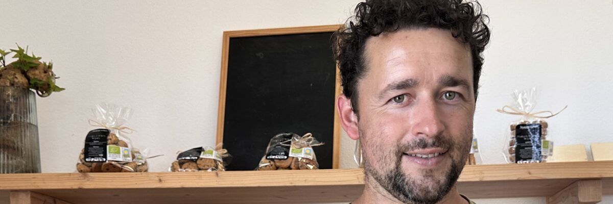 Hombre de pie en el interior de una casa, junto a una estantería con una pizarra al fondo, sonriendo a la cámara.