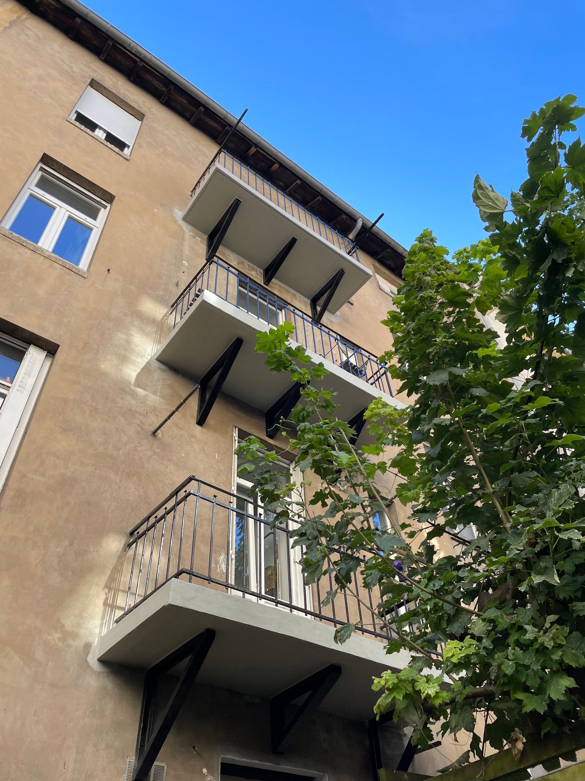 Bâtiment beige avec trois balcons, supports noirs et garde-corps blancs. Un arbre vert masque partiellement la façade.