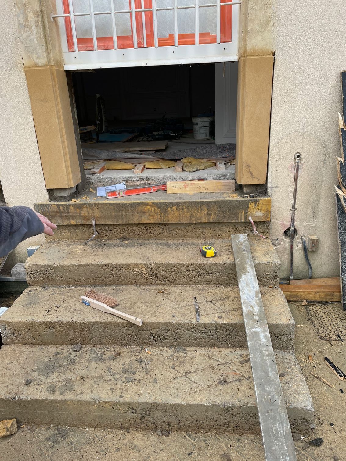 Marches en béton menant à une porte en construction ; des colonnes beiges encadrent l'ouverture.