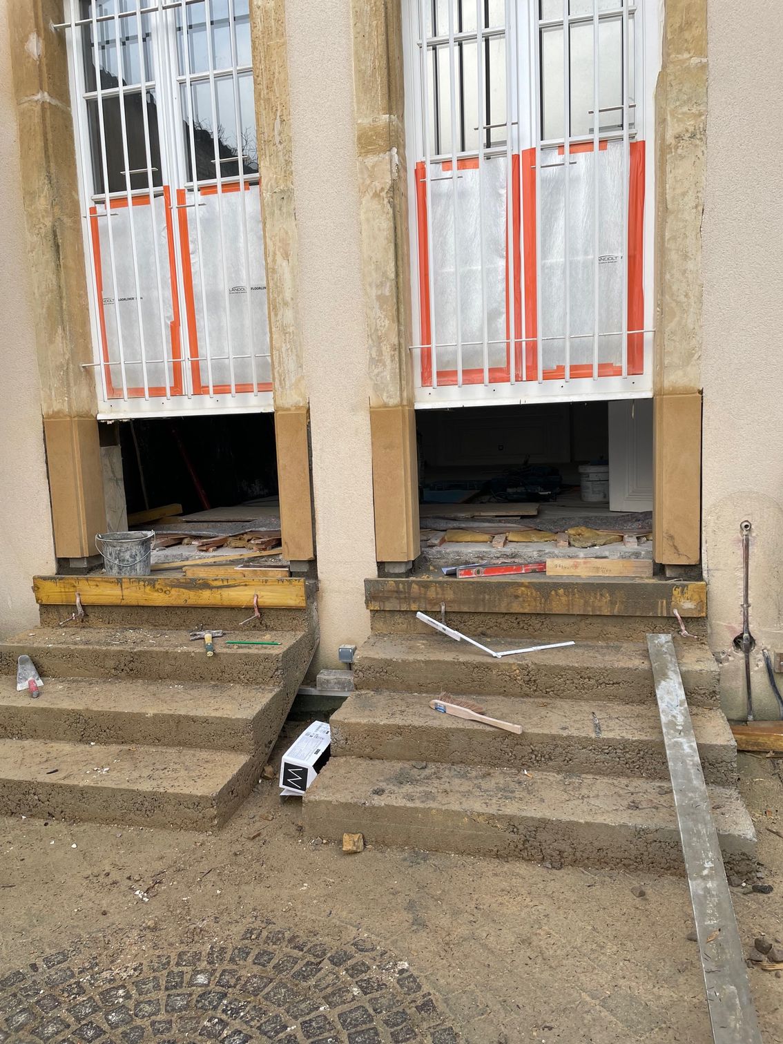 Deux châssis de fenêtres/portes en construction, marches en béton, barreaux blancs aux fenêtres, revêtement orange.