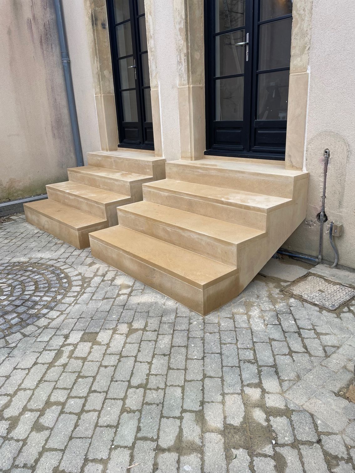 Marches en pierre menant à des portes noires dans un bâtiment, posées sur des pavés.