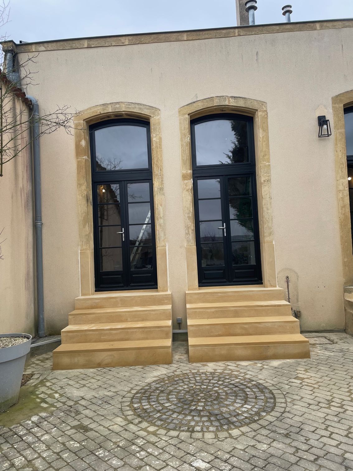 Deux portes noires avec des sommets arqués, chacune avec des marches en pierre, sur un bâtiment beige avec des accents de pierre.