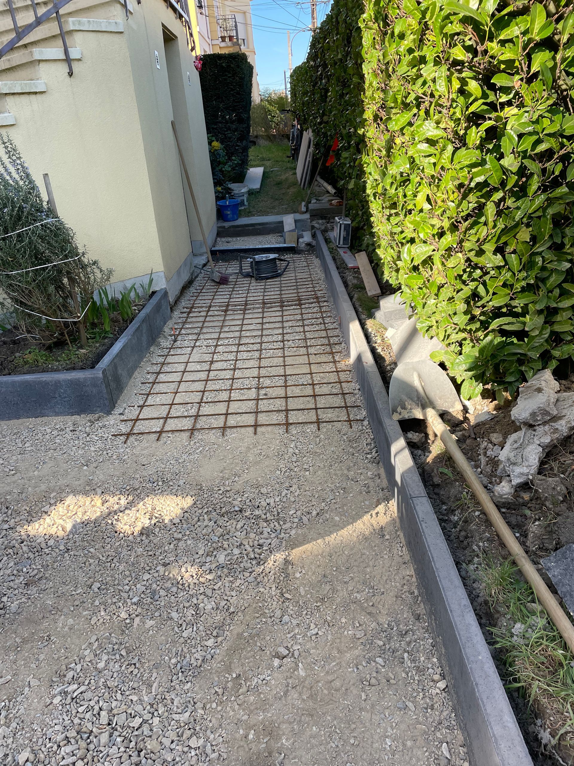 Chantier : allée en gravier avec grille en fer à béton. Bordures grises de chaque côté. Buissons verts à droite.
