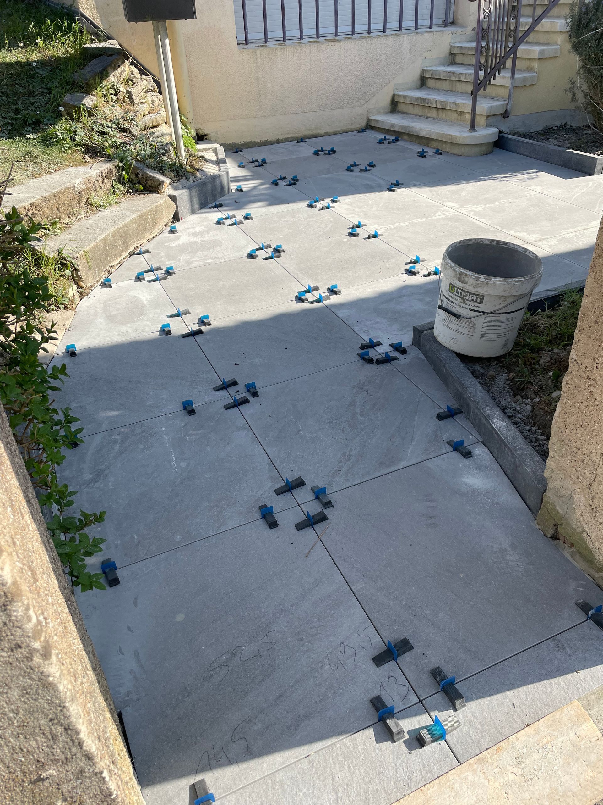 Terrasse en béton avec carrelage en place, cales bleues et seau. Lumière du soleil.