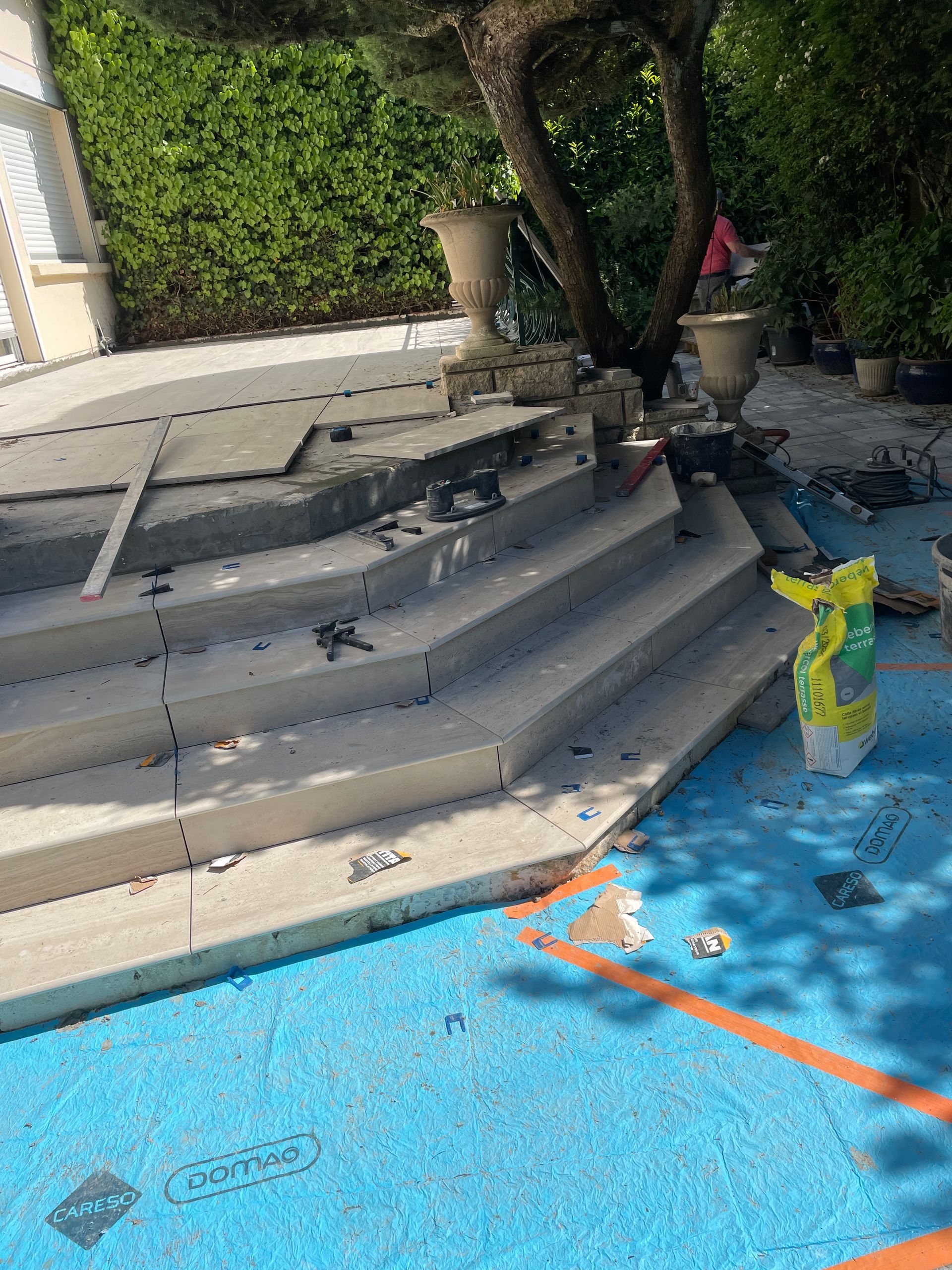 Marches en béton en construction près d'une piscine, surface bleue, feuillage vert, deux urnes visibles.