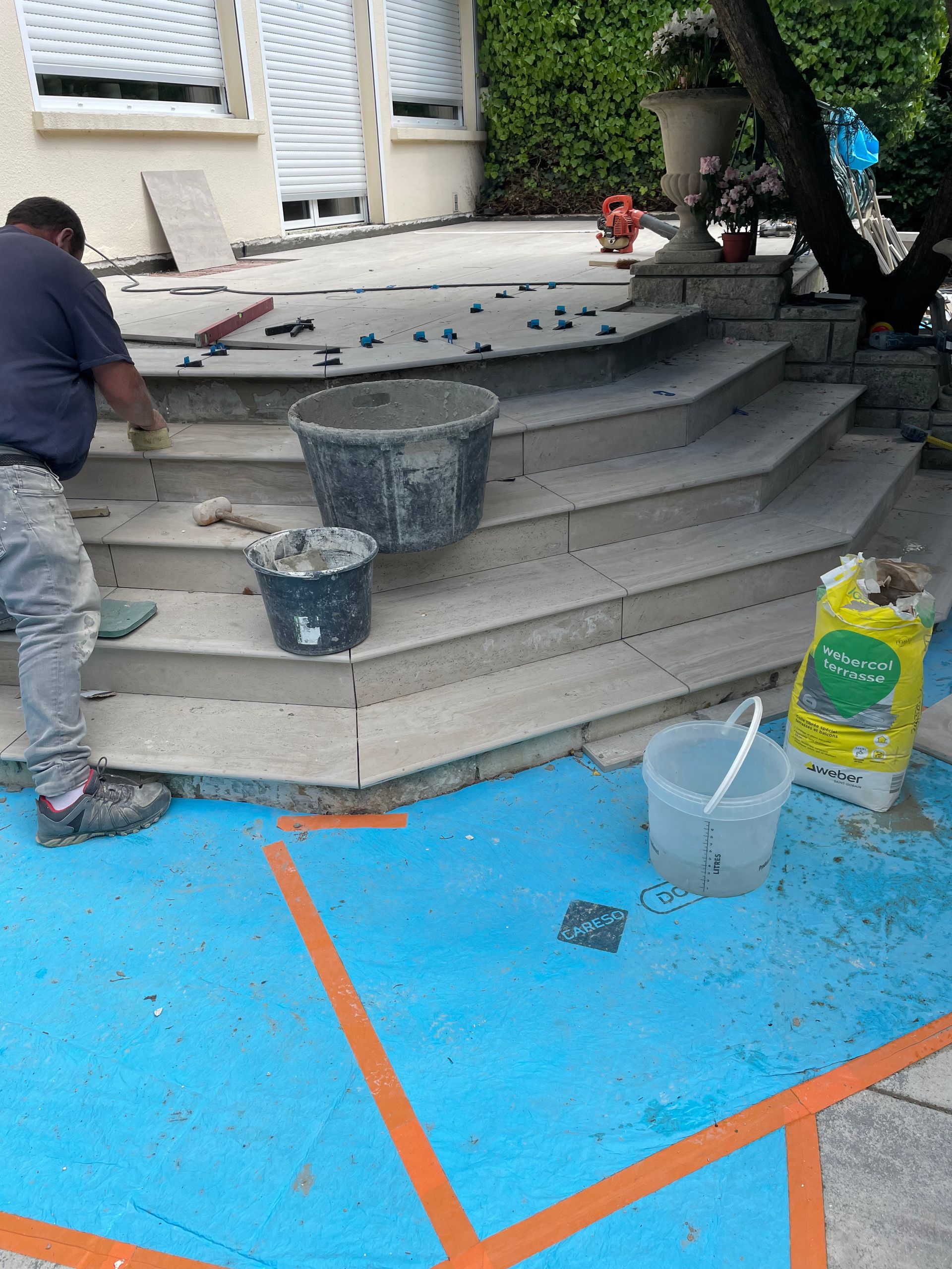 Un ouvrier carrele un escalier extérieur. Surface bleue avec lignes orange. Seaux et outils à disposition.