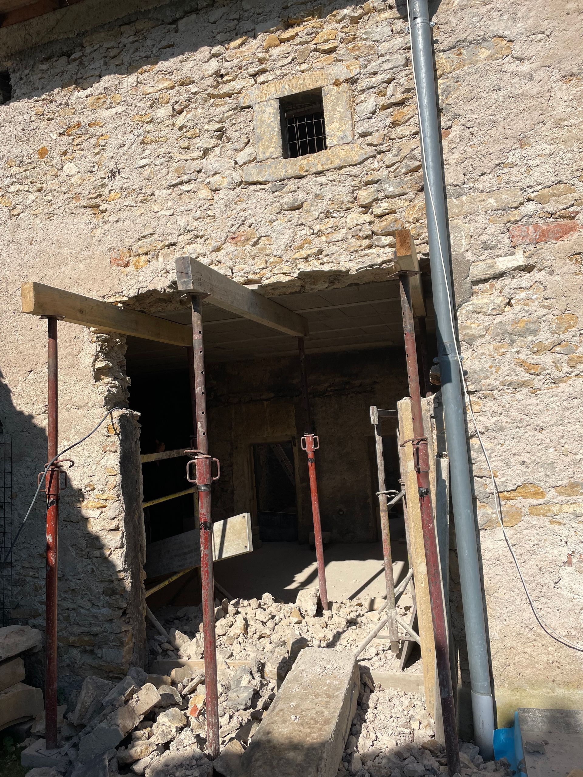 Bâtiment en pierre en construction ; ouverture de porte, soutenue par des étais métalliques ; débris au sol.