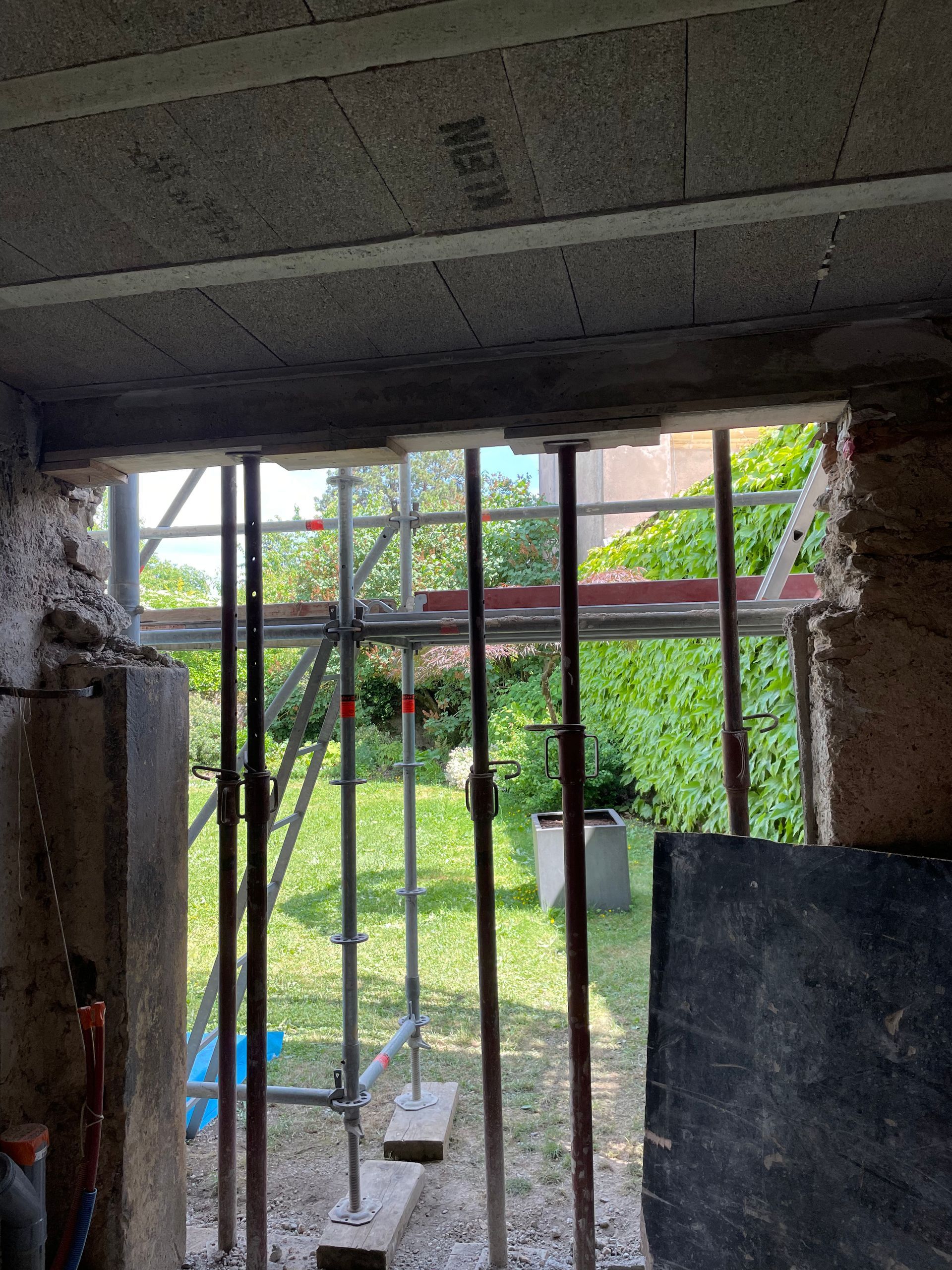 Chantier : vue intérieure d'une ouverture, soutenue par un échafaudage et des étais, avec un jardin visible à travers la fenêtre.
