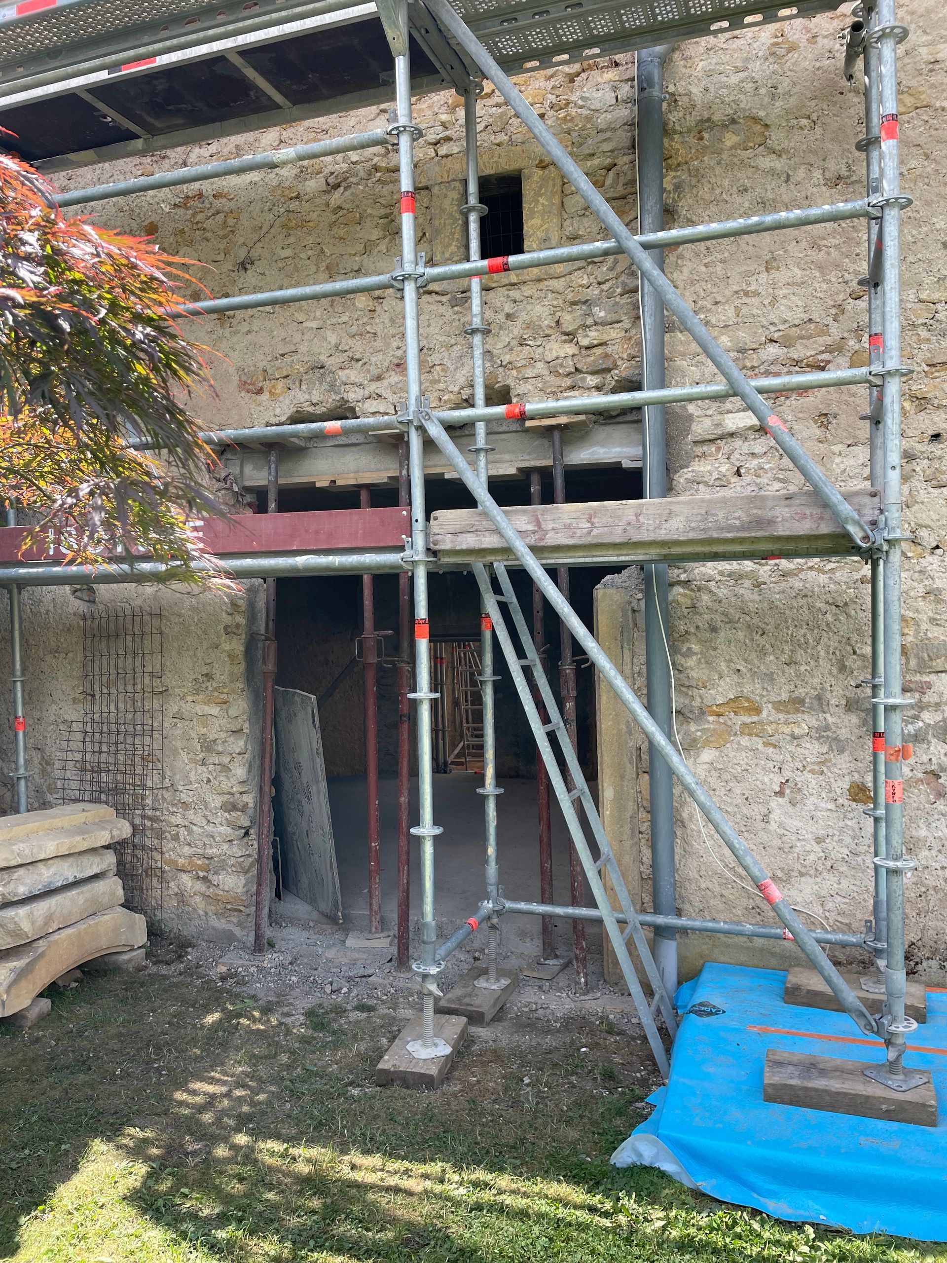 Échafaudage devant un bâtiment en pierre, entrée en cours de rénovation. Supports rouges, bâche bleue, gazon.