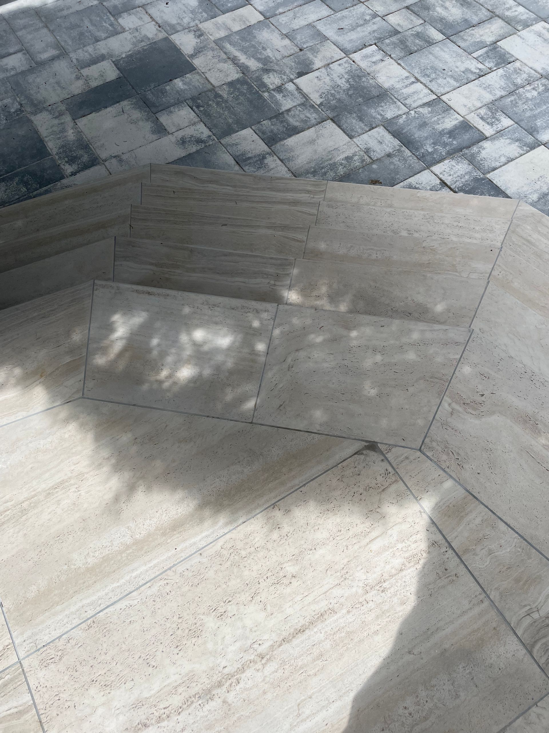 Des marches en pierre mènent à un patio en pierre à motifs, avec des ombres provenant des arbres au-dessus.