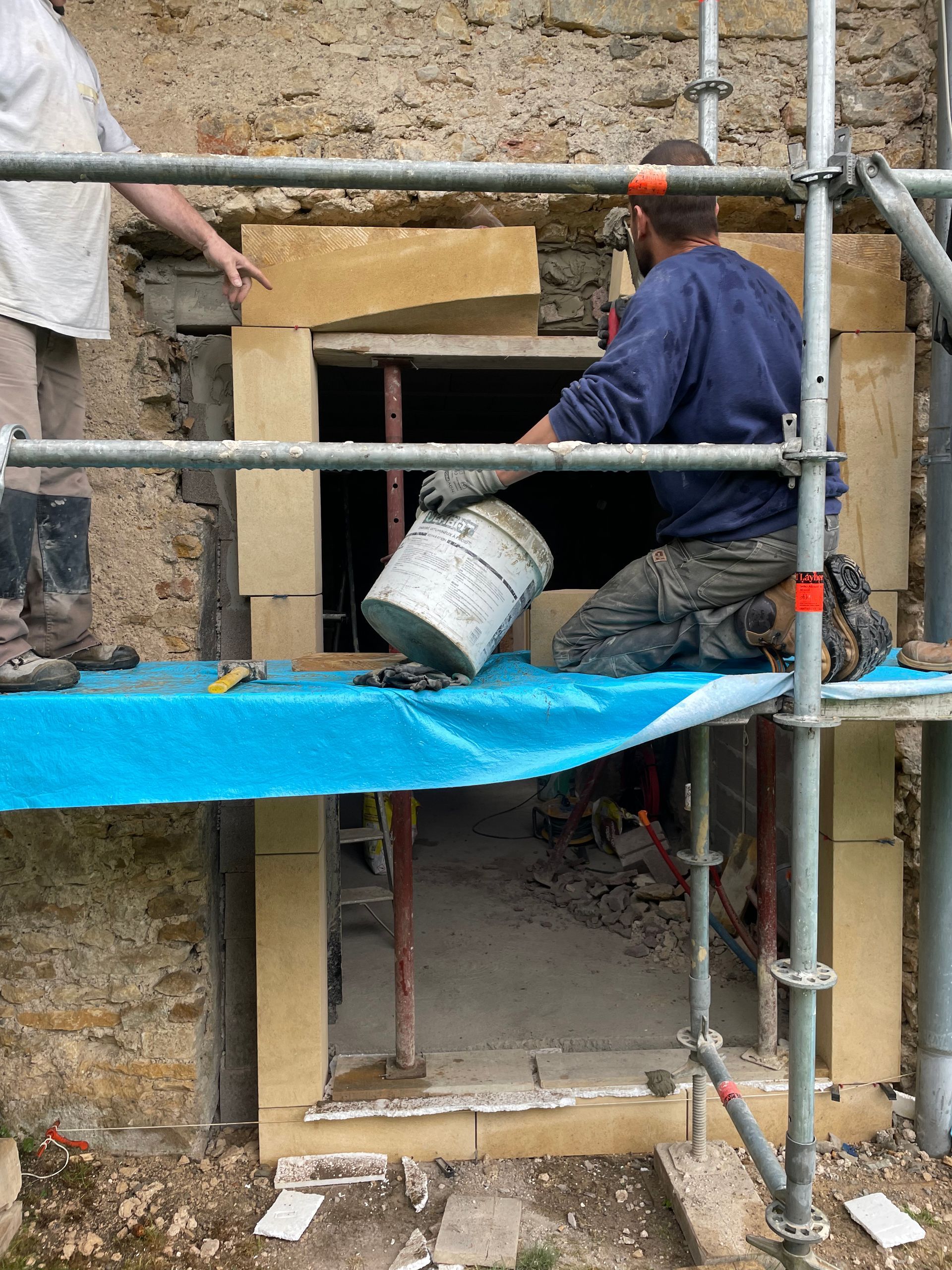 Un homme verse de l'eau d'un seau dans l'encadrement d'une porte et travaille sur la façade d'un bâtiment. Échafaudage, bâche bleue.