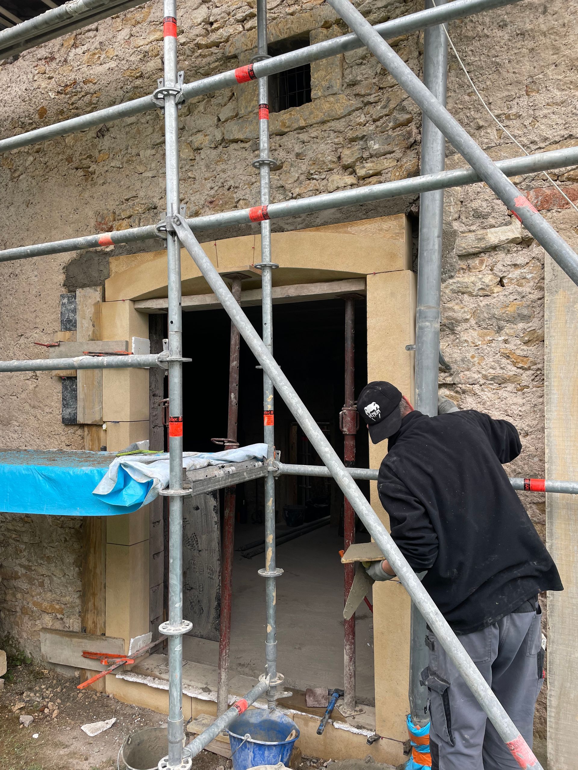 Homme travaillant sur une porte avec un échafaudage. Extérieur du bâtiment, maçonnerie beige, porte claire.