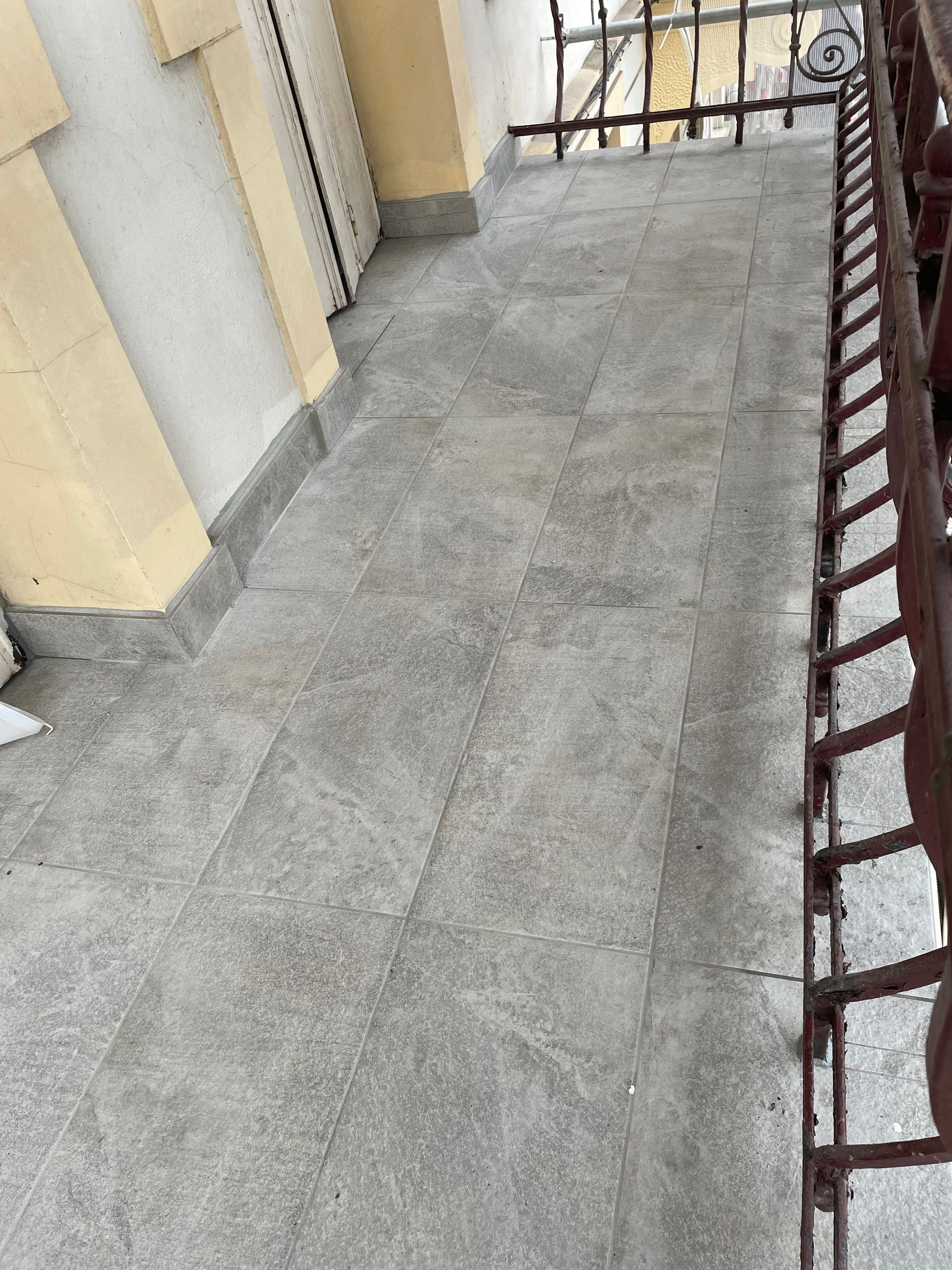 Un petit balcon avec un sol carrelé gris, une balustrade et un mur jaune.