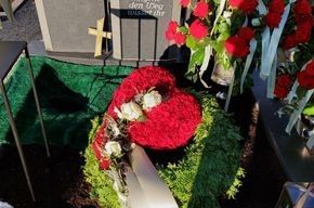 Eine Person hält einen Kranz aus roten Blumen vor einem Grab.