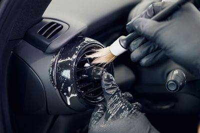 Des mains gantées nettoient la grille d'aération circulaire d'une voiture à l'aide d'une brosse et d'un chiffon.