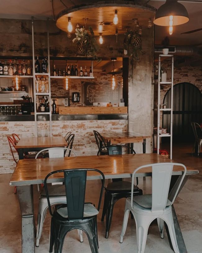 Interior de cafetería con mesas y sillas. Paredes de ladrillo visto, estanterías 