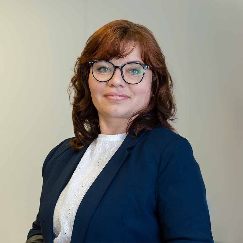 Eine Frau mit Brille und blauer Jacke steht vor einer weißen Wand.
