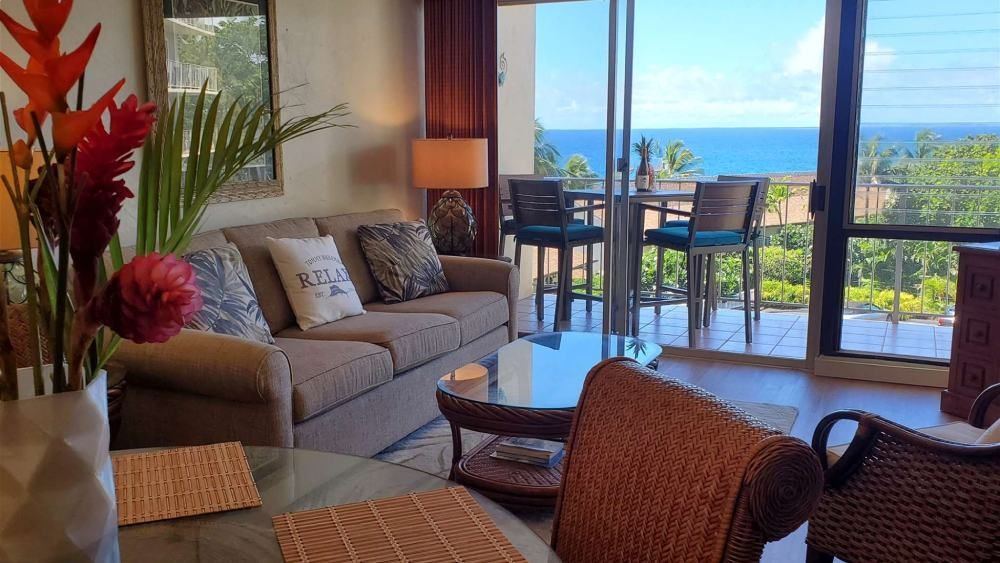 Living room with ocean view, brown sofa, table with flowers, and balcony with high-top chairs.