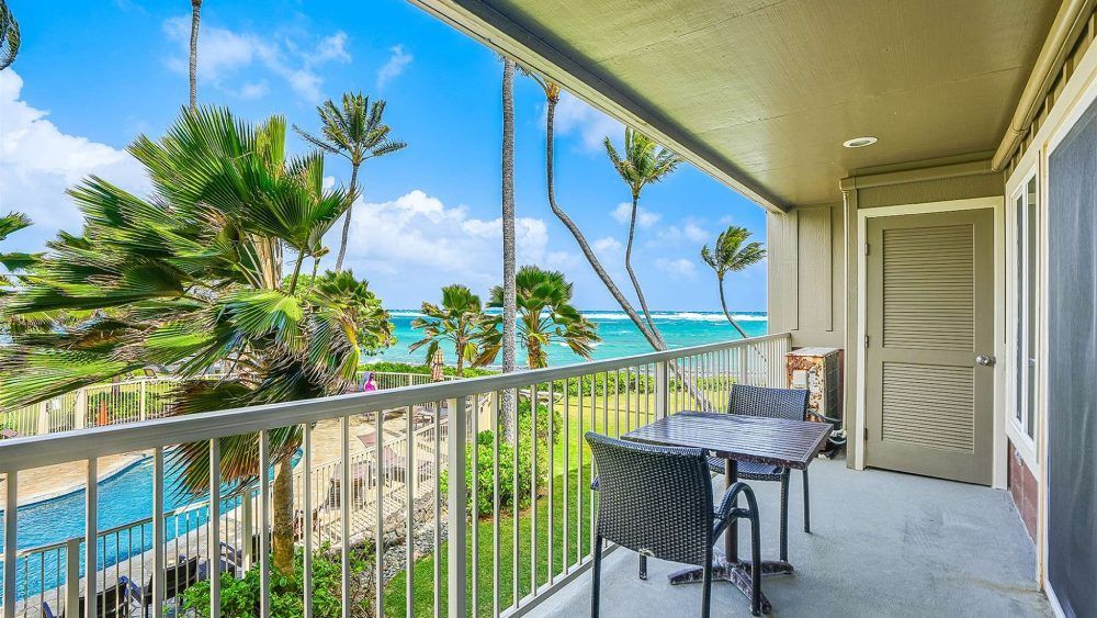 Balcony view of ocean, palm trees, and table and chairs. Sunny day.