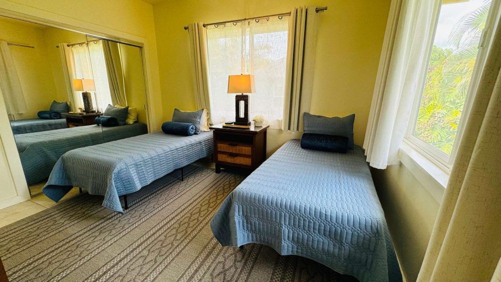 Two twin beds with blue quilts, nightstand, lamp, and window in a bedroom.