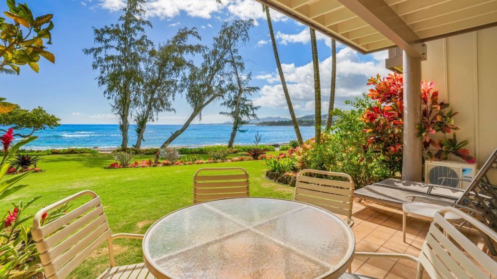 Patio with a table and chairs overlooking a grassy yard, ocean, and blue sky.