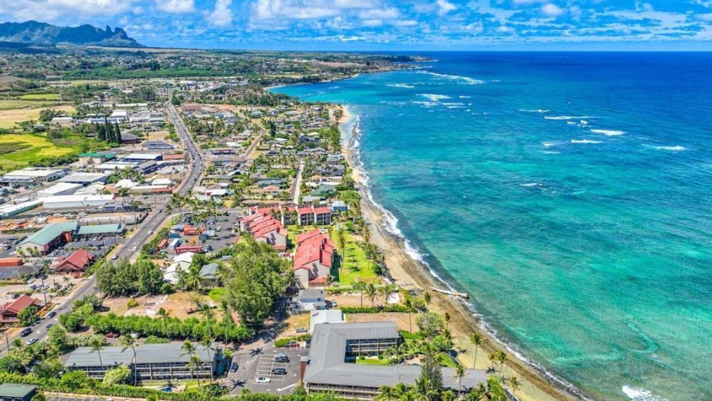Aerial view of a coastal town with buildings, a road, beach, and turquoise ocean under a blue sky.