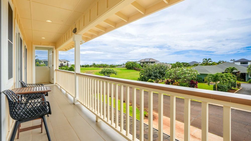 Balcony overlooking a green lawn and houses under a cloudy sky. Two chairs and a small table on the balcony.