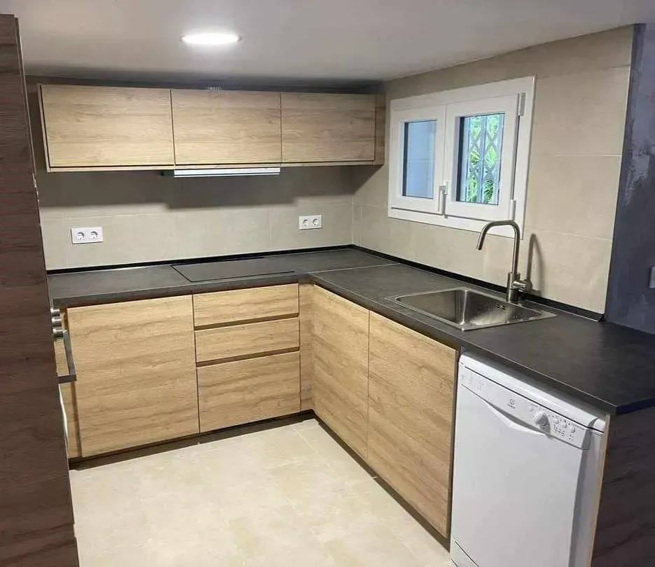 Cocina en forma de L con gabinetes de madera clara, encimeras oscuras, fregadero de acero inoxidable y lavavajillas blanco.