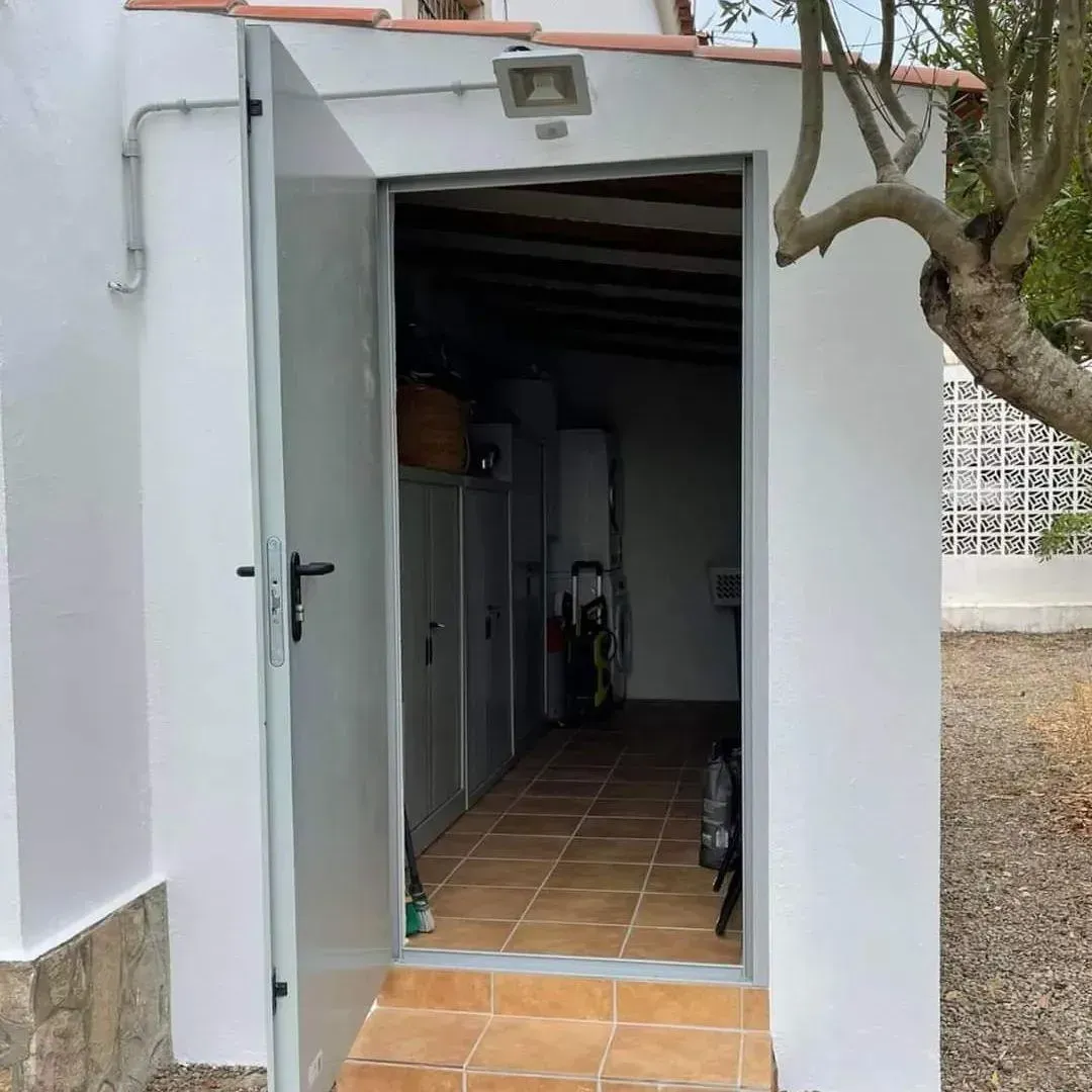 Puerta gris abierta que da a un trastero con suelo de baldosas. Paredes blancas, exterior.