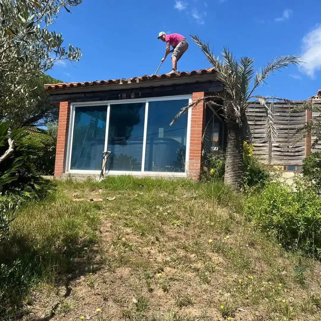 Persona en un pequeño tejado con una herramienta; un edificio con grandes ventanas y marco de ladrillo rojo, cielo azul.