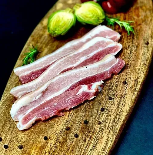 Rodanxes de panxa de porc crua amb greix marbrejat sobre una taula de fusta amb verdures i pebre.