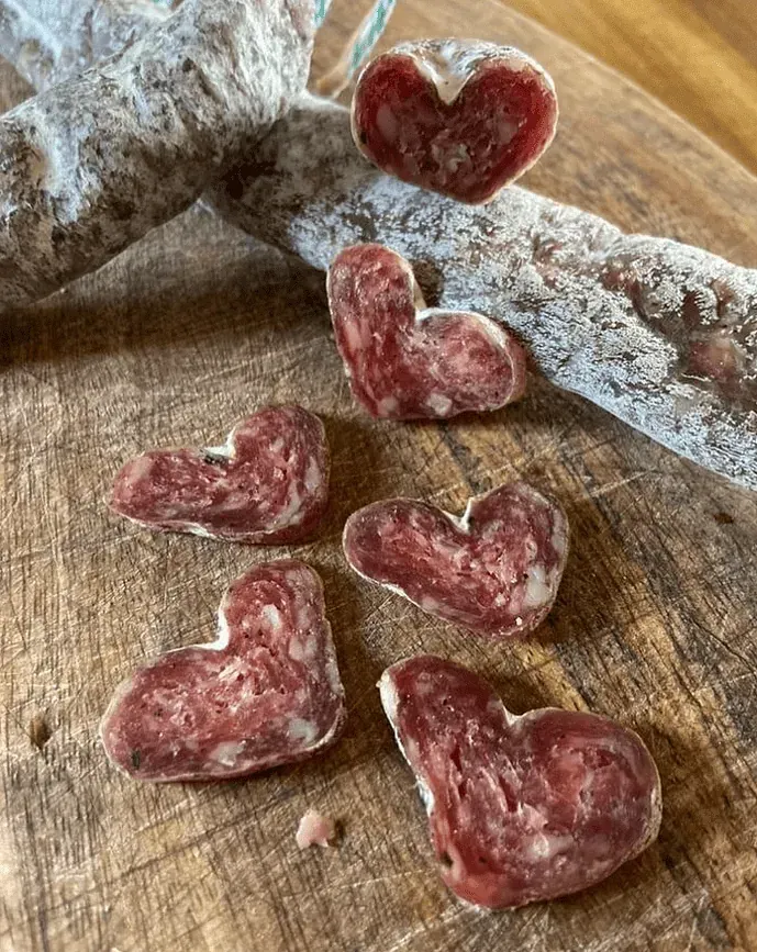 Rodanxes de salami en forma de cor sobre una superfície de fusta al costat d'una botifarra de salami.