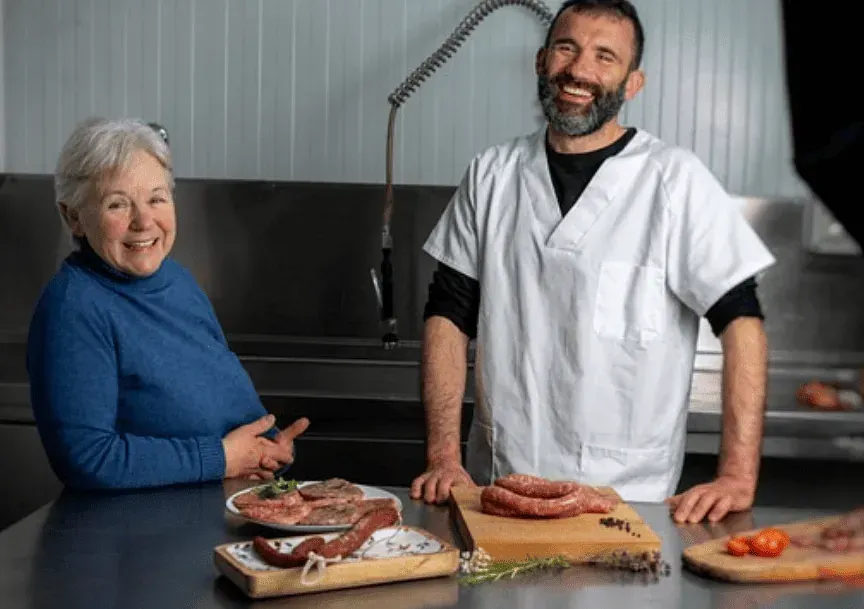 Una dona i un home somriuen al costat d'un bloc de carnisseria amb salsitxes crues, en una cuina d'acer inoxidable.