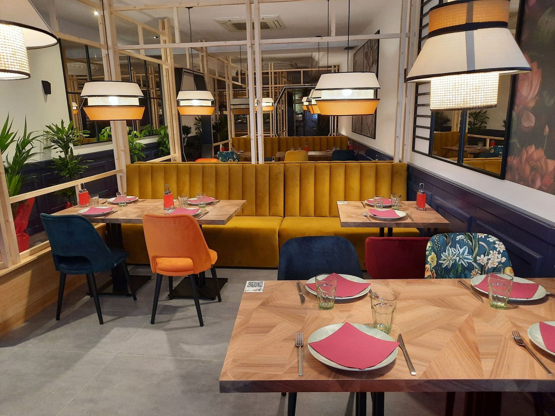 Un acogedor interior de restaurante con mesas de madera, servilletas rosas, un largo banco amarillo y coloridas sillas de terciopelo.