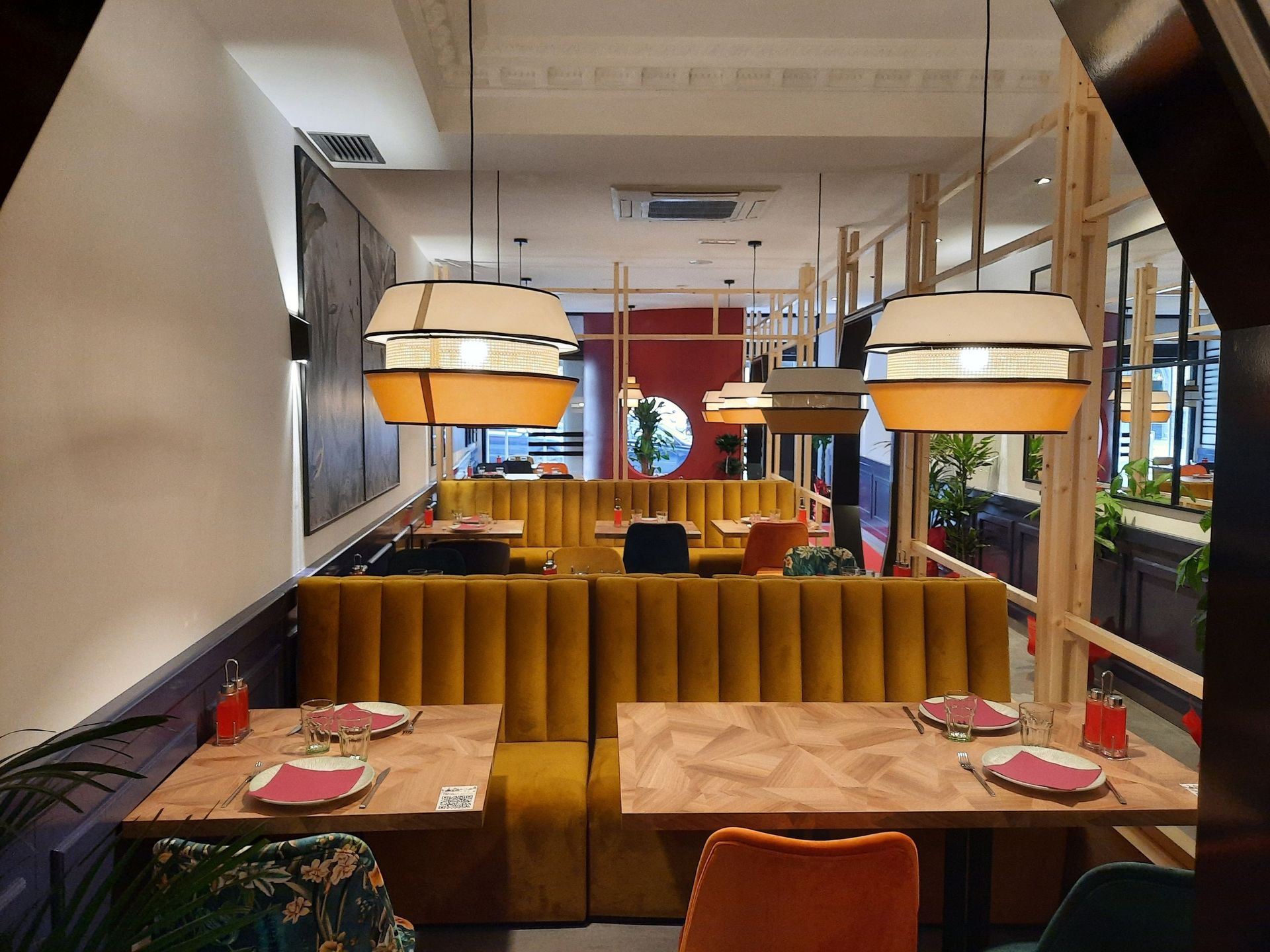 El interior de un restaurante cuenta con dos mesas de madera, cabinas tapizadas en terciopelo amarillo y grandes lámparas colgantes cilíndricas.
