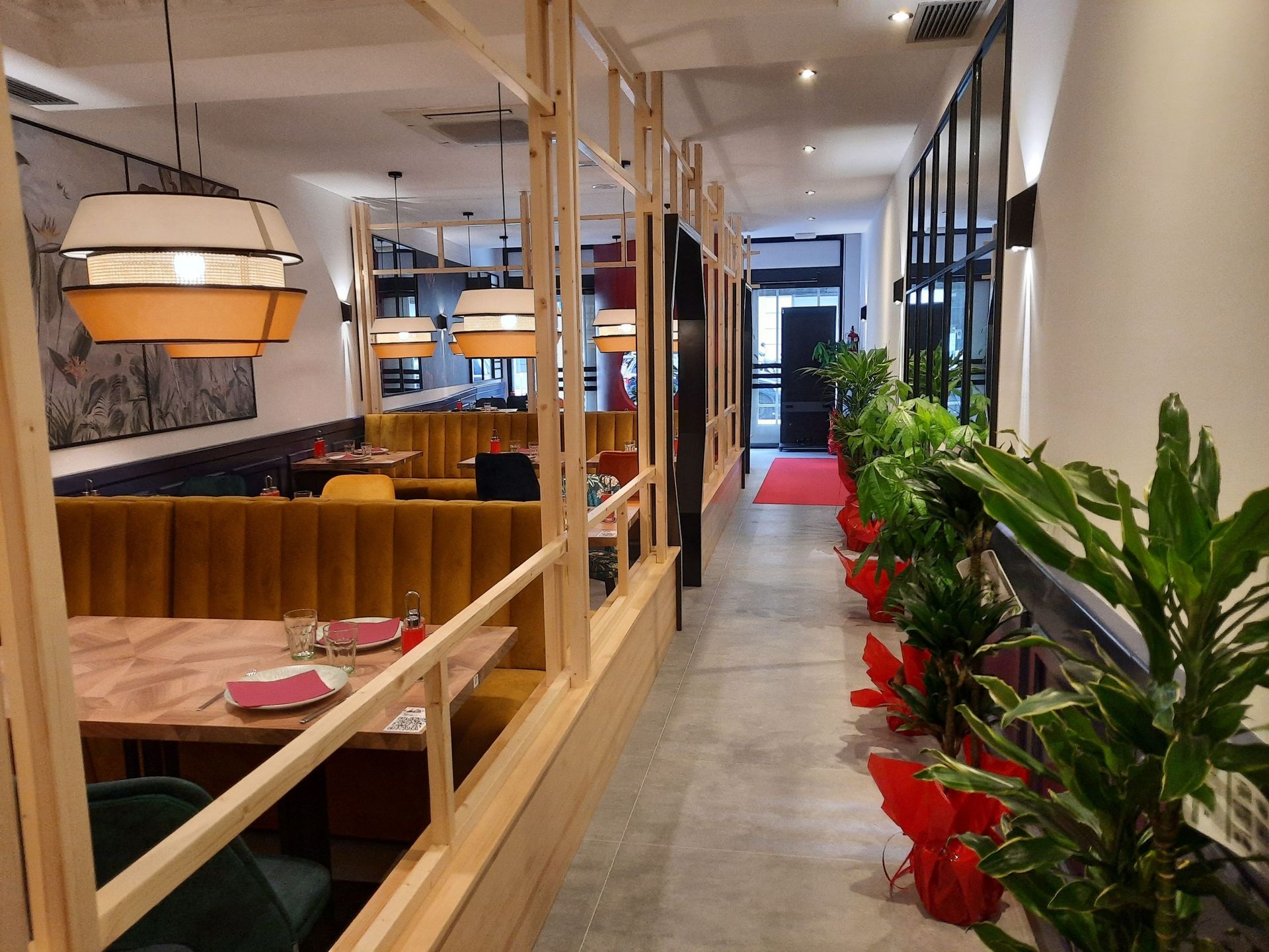 El interior de este moderno restaurante cuenta con separadores de madera, asientos tipo cabina de color amarillo, iluminación colgante y plantas en macetas a lo largo de un pasillo.