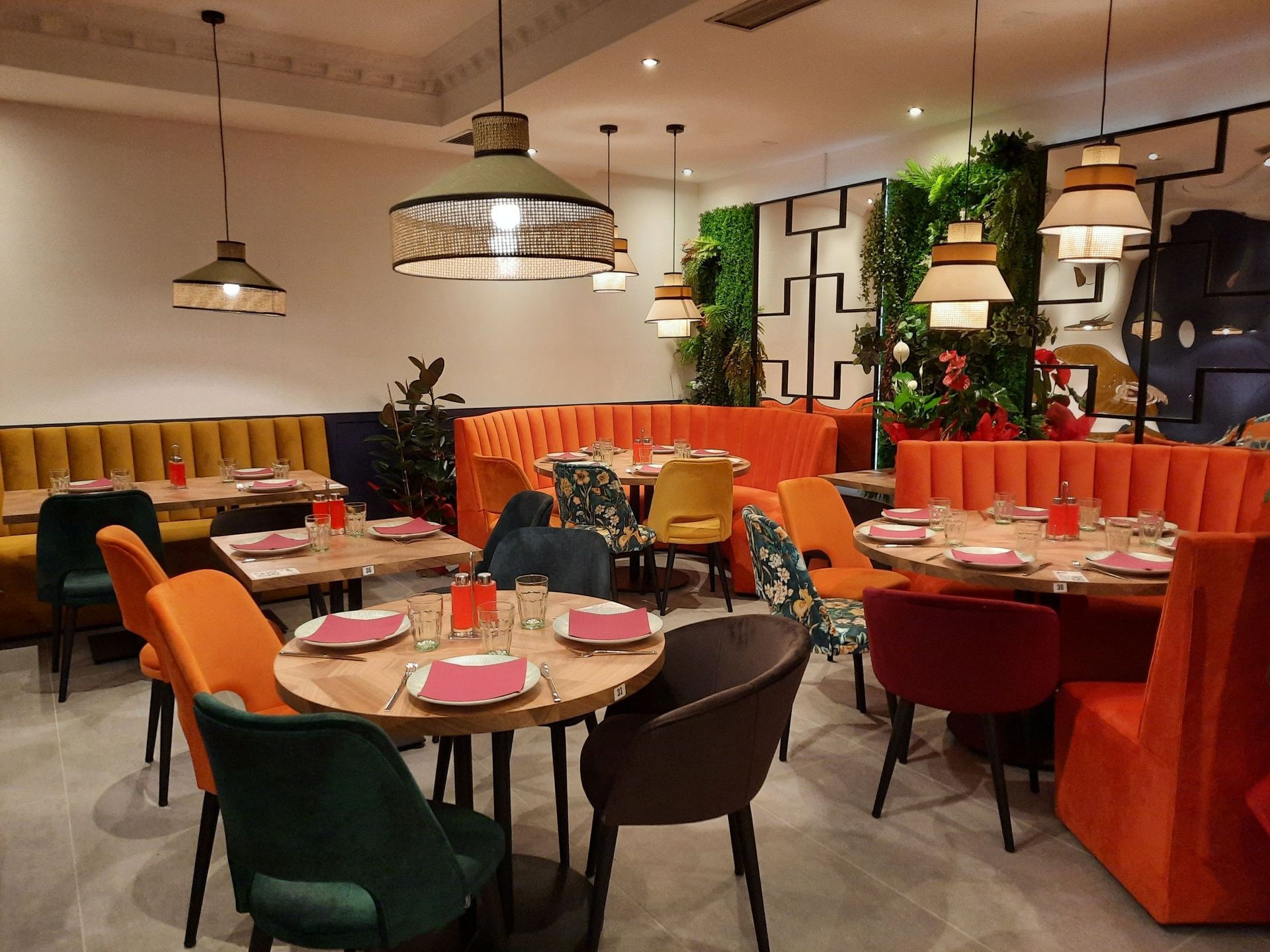 Interior de restaurante moderno con mesas redondas, cabinas de terciopelo naranja y amarillo, y sillas coloridas bajo lámparas colgantes.