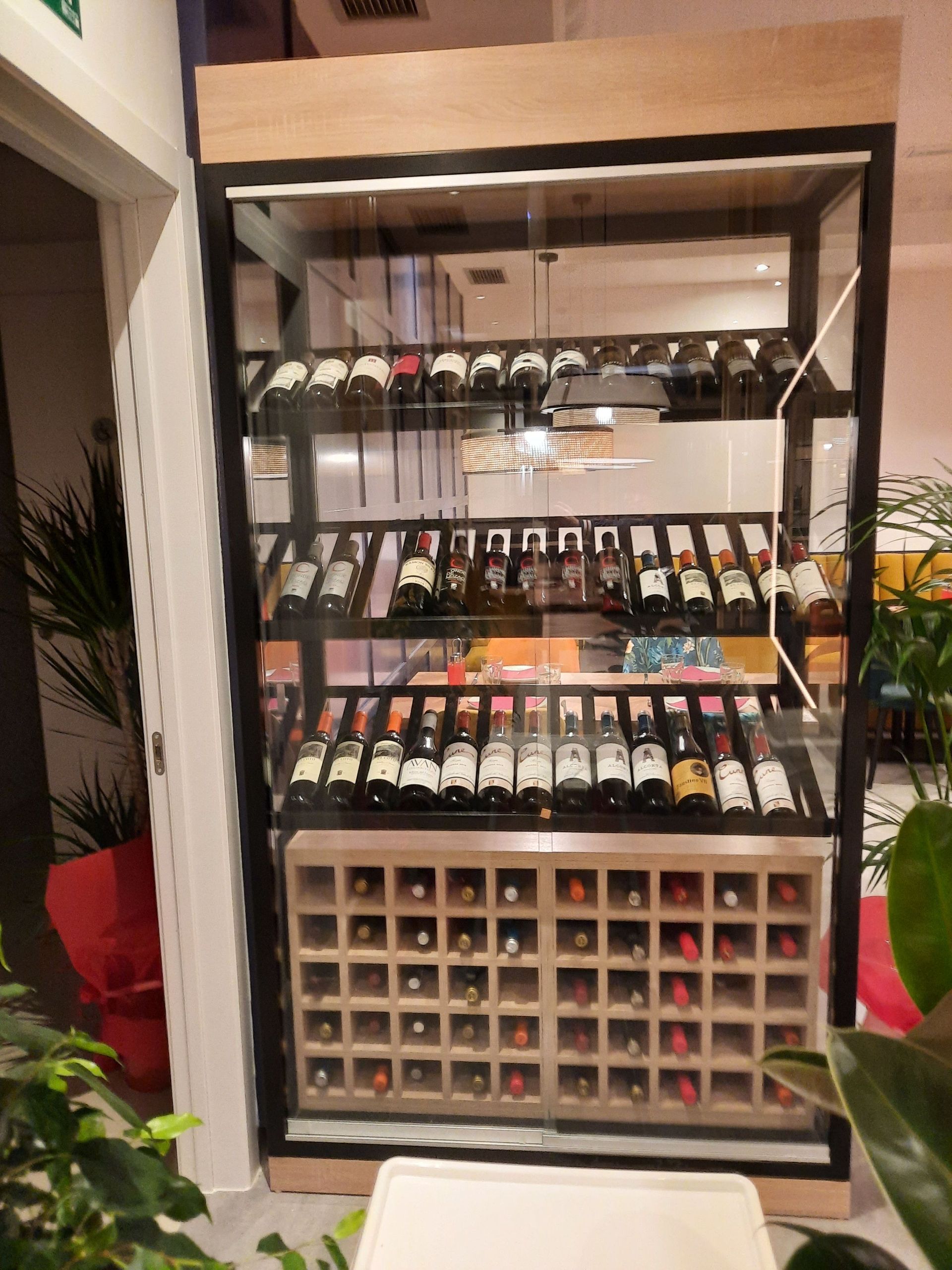 Una vitrina para vinos moderna, alta y con frente de cristal, en un restaurante, con estantes para botellas organizados y compartimentos de madera.