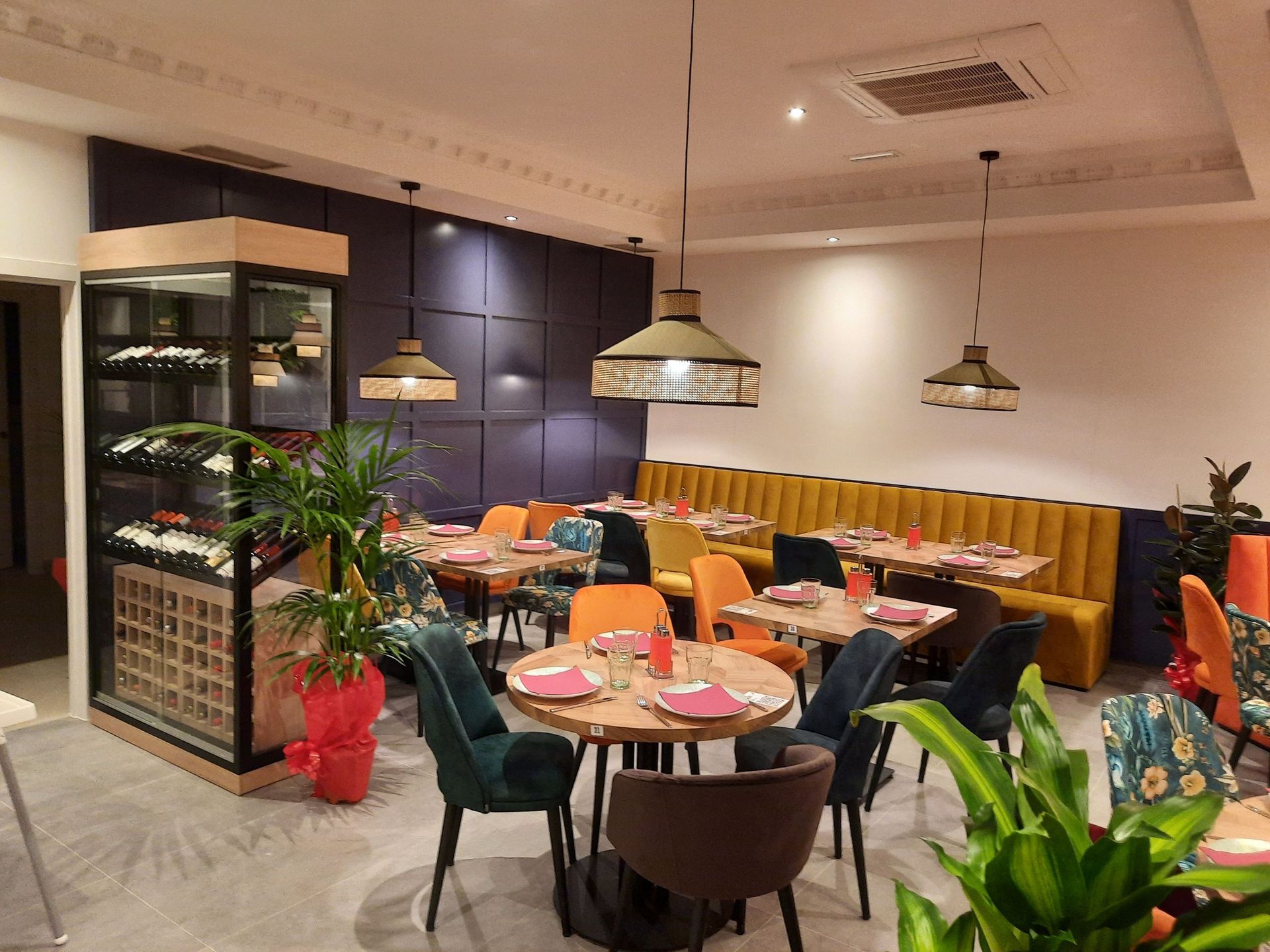 Interior de restaurante moderno con mesas redondas y cuadradas de madera, sillas de terciopelo y un banco corrido amarillo.