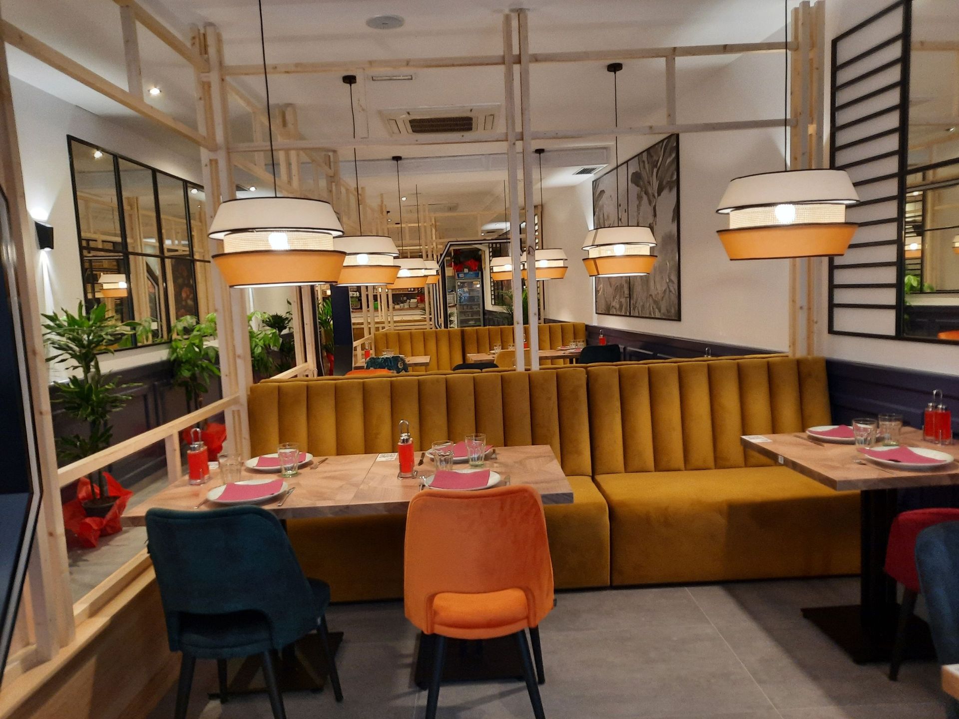 El interior del restaurante presenta mesas con sillas azules y naranjas frente a un largo reservado de terciopelo amarillo, iluminado por lámparas colgantes.