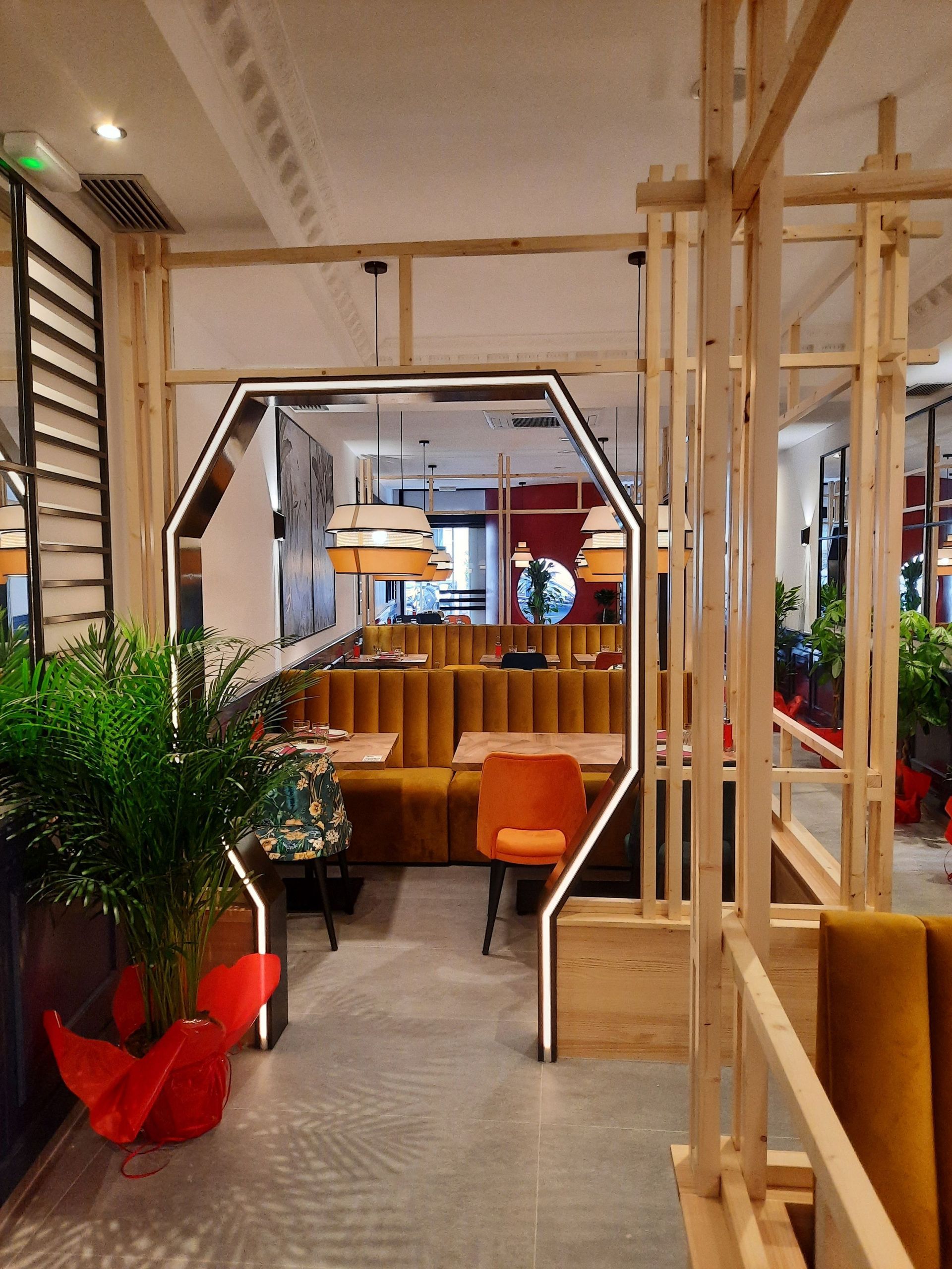 El interior de un restaurante moderno presenta estructuras de madera, arcos hexagonales luminosos y asientos tipo cabina de color amarillo mostaza.