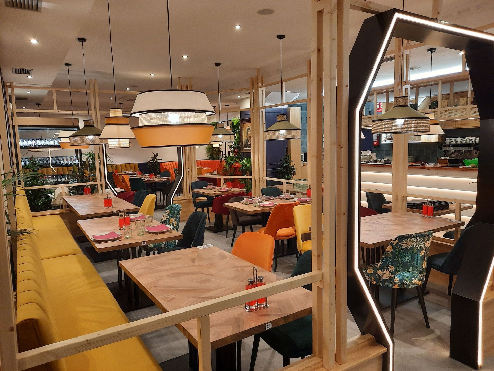 Un moderno comedor de restaurante con mesas de madera, bancos amarillos, sillas coloridas y lámparas colgantes cálidas de diseño geométrico.