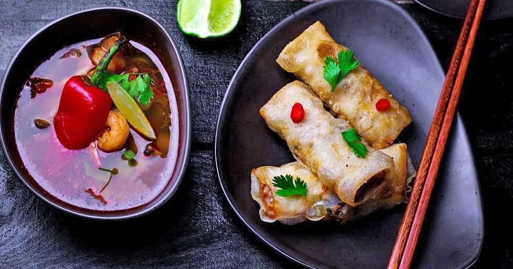 Un tazón oscuro de sopa con lima y chile reposa junto a un plato de rollitos de primavera crujientes servidos con palillos.