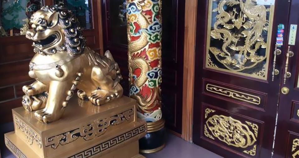 Una estatua dorada de un león guardián chino sobre un pedestal, junto a un pilar decorativo con forma de dragón y una puerta ornamentada con motivos dorados.