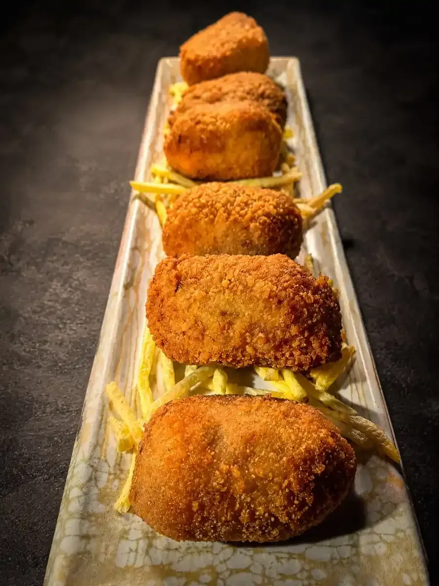 Croquetas empanizadas fritas sobre papas fritas servidas en una bandeja blanca larga, vista de cerca.