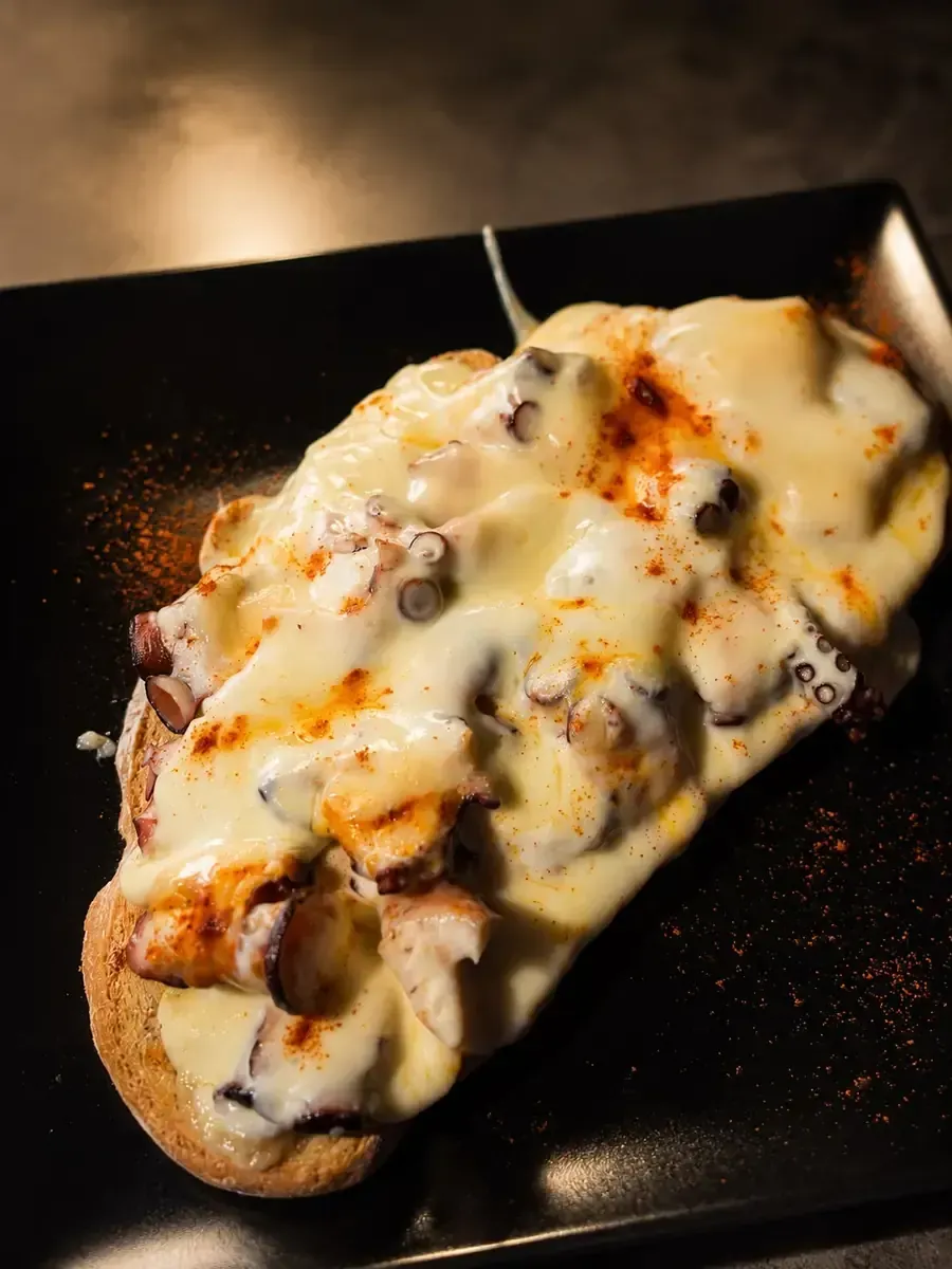Pan horneado con queso, cubierto con queso derretido y champiñones en un plato negro.