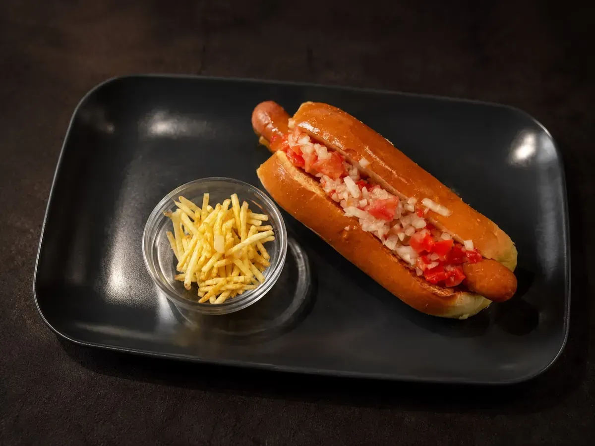 Perrito caliente con kétchup y cebolla en un plato negro, con un pequeño cuenco de queso rallado al lado.