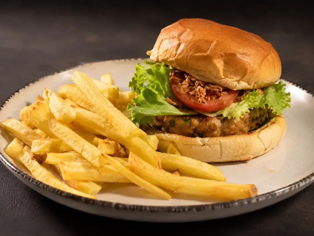 Hamburguesa y patatas fritas en un plato blanco, con lechuga y tomate sobre una mesa oscura.